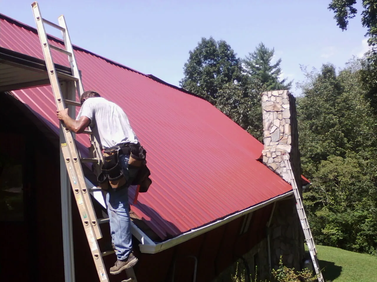 Roofer on a ladder beside a red metal roof and stone chimney — Soffit & Fascia Repair by Roofing Brothers in Taopi